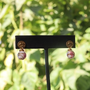 May include: A pair of earrings with a gold-colored hammered disc top and a dangling, round, glass bead. The glass bead is white with pink and gold floral designs. The earrings are displayed on a black stand against a blurred green background.