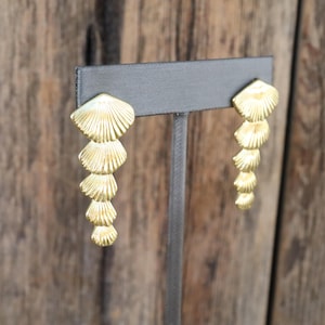 Könnte beinhalten: Goldfarbene Hängeohrringe mit Muscheldesign. Jeder Ohrring hat eine Reihe von abgestuften, fächerförmigen Elementen, die einen Kaskadeneffekt erzeugen. Die Ohrringe werden auf einem grauen Ständer vor einem Holzgrund gezeigt.
