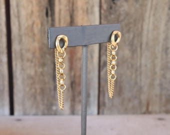 Op de afbeelding: Een paar goudkleurige oorbellen met een kleine ovale knop verbonden aan een verticale reeks van drie ronde schakels en een fijne bungelende ketting. De oorbellen worden gepresenteerd op een donkergrijze T-vormige standaard voor weergave.