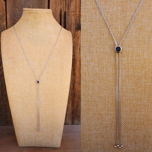 Puede incluir: Un collar tipo lariat plateado con un colgante circular azul oscuro. El collar presenta una larga cadena colgante con tres pequeñas cuentas redondas plateadas en el extremo. El collar se exhibe en un maniquí beige.