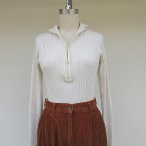 May include: A white ribbed sweater with a zipper up the front and long sleeves. The sweater is paired with brown corduroy pants.