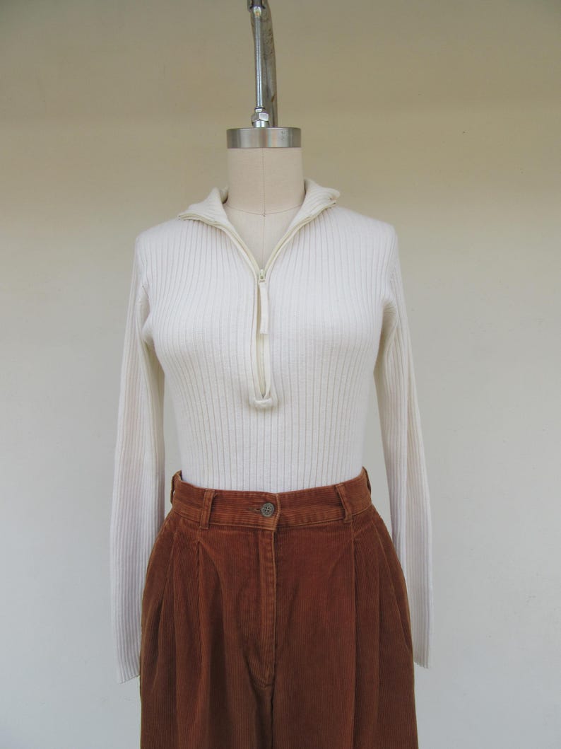May include: A white ribbed sweater with a zipper up the front and long sleeves. The sweater is paired with brown corduroy pants.