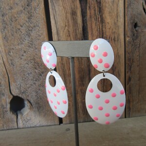 May include: A pair of white oval earrings with pink polka dots. The earrings are dangling and have a small circle cut out of the bottom of the larger oval.