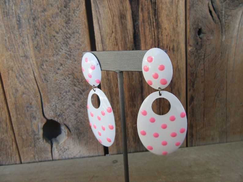 May include: A pair of white oval earrings with pink polka dots. The earrings are dangling and have a small circle cut out of the bottom of the larger oval.