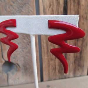 May include: A pair of red lightning bolt shaped earrings on a white display stand.