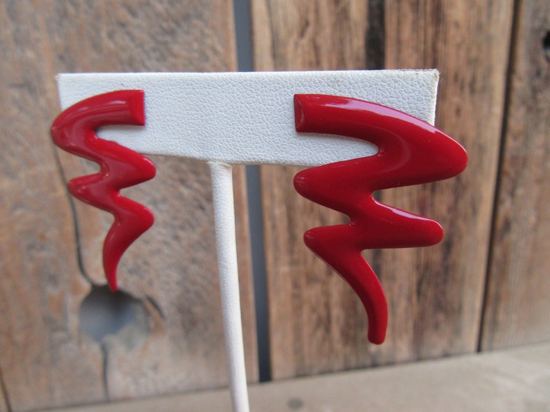 May include: A pair of red lightning bolt shaped earrings on a white display stand.