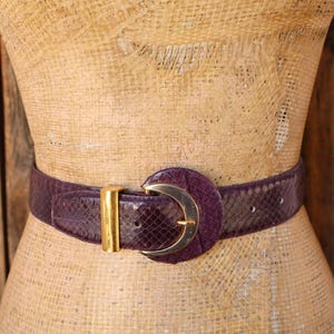 Può includere: Una cintura viola scuro con un motivo testurizzato a pelle di serpente, caratterizzata da una grande fibbia rotonda viola scuro con un accento metallico argentato a forma di mezzaluna e un passante rettangolare color oro.