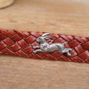 May include: Close-up of a brown leather belt with a woven design. A silver-coloured metal rabbit charm is attached to the belt. The belt is a warm reddish-brown colour.