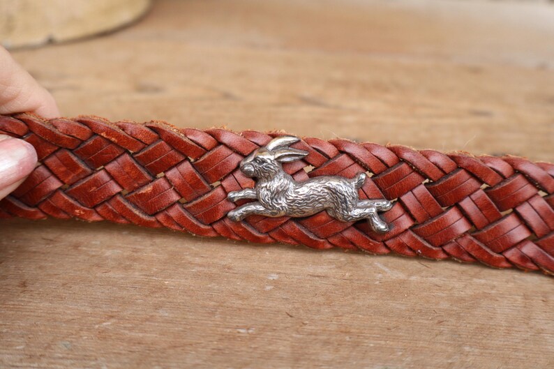 May include: Close-up of a brown leather belt with a woven design. A silver-coloured metal rabbit charm is attached to the belt. The belt is a warm reddish-brown colour.