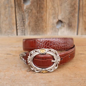 May include: A brown leather belt with a textured, crocodile-like pattern. The belt features a silver-toned buckle with an oval shape and decorative gold accents. The belt is displayed on a wooden surface.