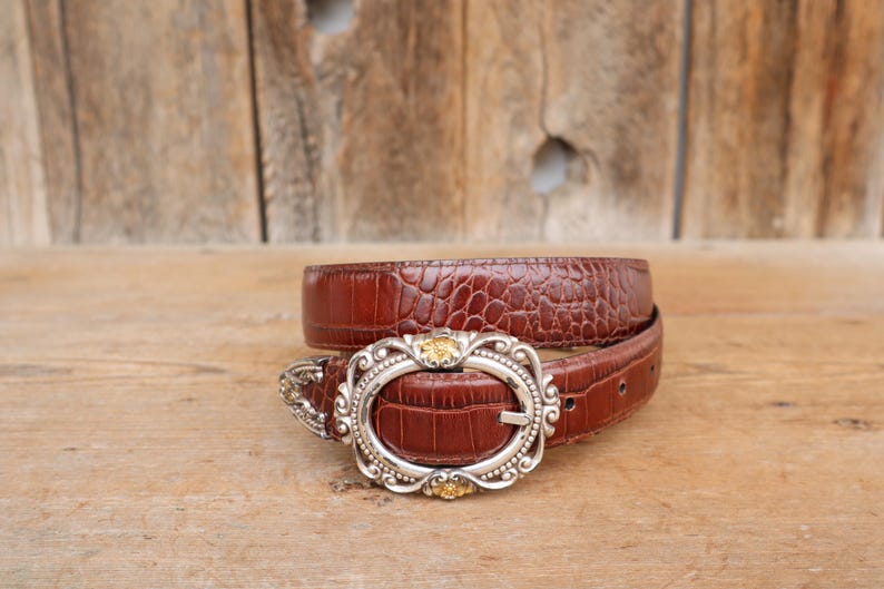May include: A brown leather belt with a textured, crocodile-like pattern. The belt features a silver-toned buckle with an oval shape and decorative gold accents. The belt is displayed on a wooden surface.