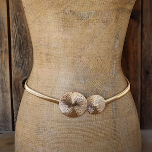 May include: Gold-toned metal belt with two circular, textured medallions. The belt is displayed on a mannequin, showcasing its design and fit. The medallions have a sunburst pattern, adding a decorative element to the accessory.