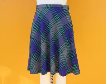May include: A vintage plaid skirt in shades of purple, teal, and gray. The skirt features a classic plaid pattern with a button closure and a flared silhouette. The skirt is displayed on a mannequin against a yellow background.