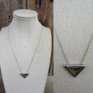 May include: A silver-toned necklace featuring a triangular pendant. The pendant has a polished, reflective surface. The necklace is displayed on a mannequin, showcasing its minimalist design. The chain is delicate, and the pendant is approximately 1 inch wide.