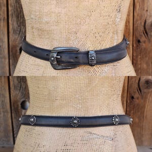 May include: Two black leather belts with silver metal detailing. The top belt has a decorative buckle and loop. The bottom belt features floral studs. Both belts are shown on a mannequin against a wooden backdrop.