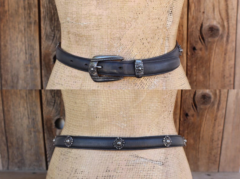 May include: Two black leather belts with silver metal detailing. The top belt has a decorative buckle and loop. The bottom belt features floral studs. Both belts are shown on a mannequin against a wooden backdrop.