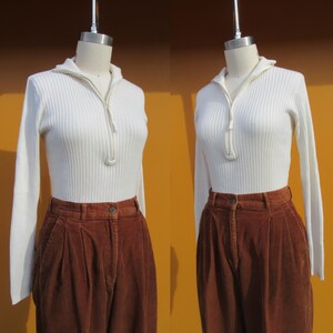 May include: A white ribbed sweater with a zipper at the neck and long sleeves. The sweater is paired with brown corduroy pants.