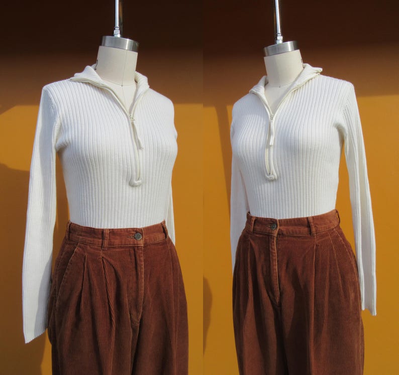 May include: A white ribbed sweater with a zipper at the neck and long sleeves. The sweater is paired with brown corduroy pants.