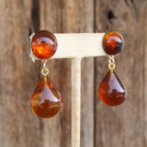 May include: A pair of amber-colored drop earrings. Each earring features a round stud and a teardrop-shaped pendant. The earrings are connected by a gold-colored wire. The earrings are displayed on a beige stand against a wooden background.