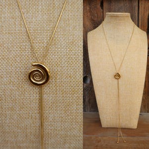 May include: A gold-colored lariat necklace featuring a spiral pendant. The necklace has a delicate chain that extends down, ending with two small, round accents. The pendant is a detailed spiral design.