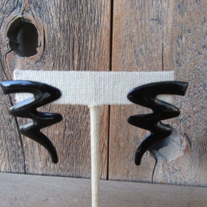 Pode incluir: Um par de brincos pretos de forma abstrata. Os brincos s&atilde;o feitos de um material liso e brilhante e t&ecirc;m um design geom&eacute;trico.