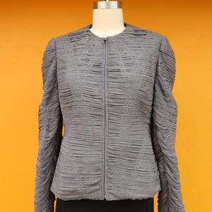 May include: A gray jacket with a textured, pleated design. The jacket has a zipper closure and long sleeves with a gathered, puffy design. The jacket is displayed on a mannequin against an orange background.