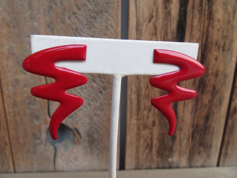 May include: A pair of red, abstract shaped earrings. The earrings are made of a shiny, smooth material and are shaped like a lightning bolt.