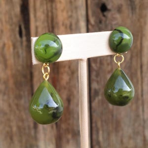 Può includere: Orecchini a clip verde oliva con effetto marmorizzato. Gli orecchini presentano una parte superiore rotonda e una inferiore a forma di goccia, collegate da un anello color oro. Lo sfondo è una superficie di legno sfocata.