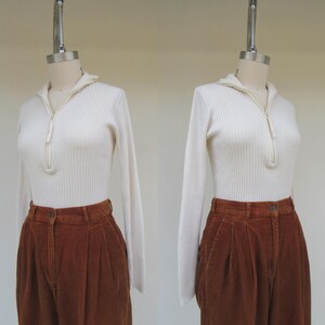 May include: A white ribbed sweater with a zipper neckline and long sleeves, paired with brown corduroy pants.