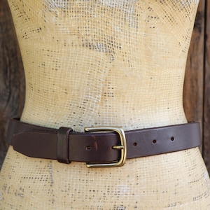 May include: A brown leather belt with a gold-colored buckle is displayed on a mannequin. The belt has a classic design with a rectangular buckle and several holes for adjustment. The mannequin is set against a wooden background.