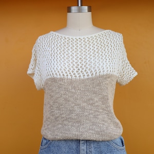 May include: A two-tone knit top with a unique design. The top features a cream-colored, open-weave upper section and a tan, close-knit lower section. The short-sleeved shirt is displayed on a mannequin against an orange background.