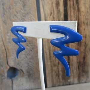 May include: A pair of blue abstract shaped earrings on a white display stand. The earrings are made of a smooth, glossy material.