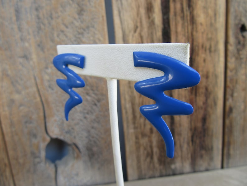 May include: A pair of blue abstract shaped earrings on a white display stand. The earrings are made of a smooth, glossy material.