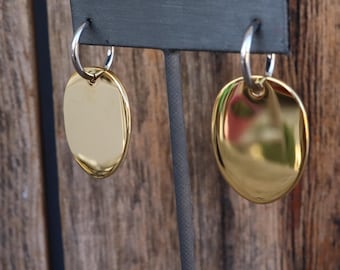 Op de afbeelding: Een paar gouden ovale oorbellen met een zilveren hoepel. De oorbellen worden getoond op een zwarte standaard.