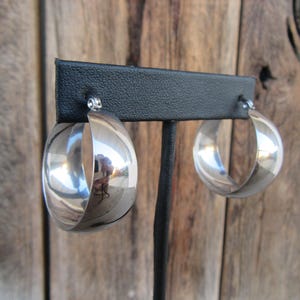 May include: A pair of polished silver hoop earrings. The earrings are displayed on a black stand against a wooden background. The hoops have a rounded, open design, reflecting light and the surrounding environment.