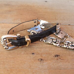 May include: A vintage black leather belt with a decorative silver-tone buckle and ornate metal accents. The belt features a gold-tone clasp and a light brown leather backing. The belt is lying on a wooden surface.