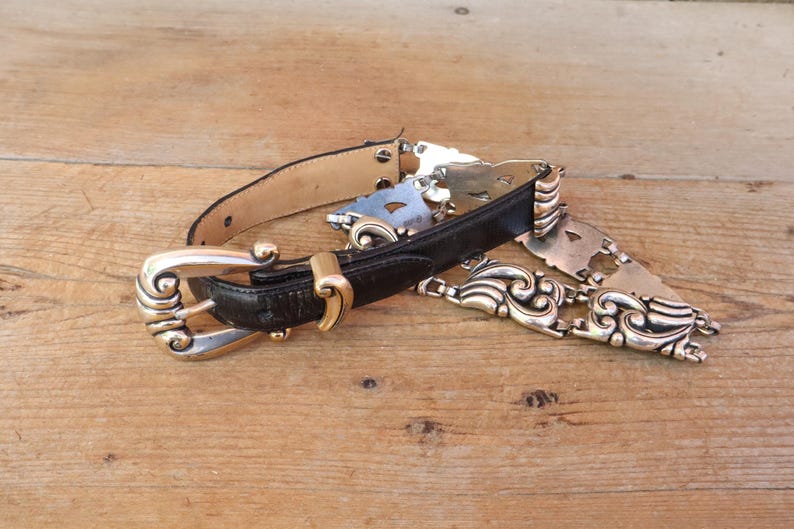 May include: A vintage black leather belt with a decorative silver-tone buckle and ornate metal accents. The belt features a gold-tone clasp and a light brown leather backing. The belt is lying on a wooden surface.