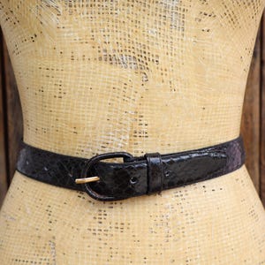 May include: A black belt with a snakeskin pattern is fastened around a mannequin's waist. The belt has a gold-colored buckle. The mannequin is tan and the background is a wooden wall.