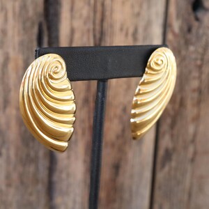 May include: A pair of gold-toned earrings with a shell-like design. Each earring features a spiral at the top and a series of curved ridges, creating a textured, eye-catching appearance. The earrings are displayed on a black stand against a wooden background.