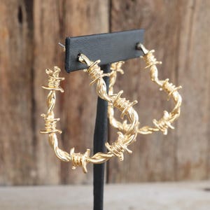 May include: Gold-colored hoop earrings with a barbed wire design. The earrings are displayed on a black stand against a wooden background. The hoops are approximately 2 inches in diameter.
