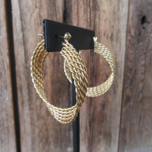 May include: Gold-coloured hoop earrings with a woven design. The earrings are displayed on a black stand against a wooden background. The hoops have a textured, braided appearance, catching the light and adding a touch of elegance to the jewellery.