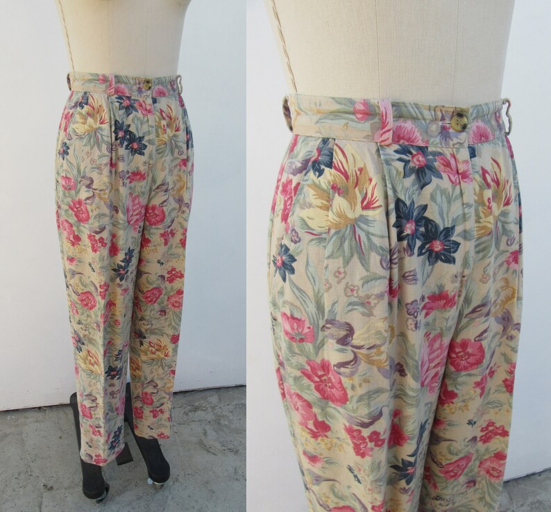 May include: Beige high-waisted pants with a floral print featuring pink, yellow, and blue flowers. The pants have a pleated front and a wide waistband.