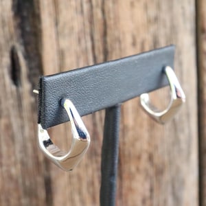 May include: A pair of polished silver earrings with a geometric, open design. The earrings are displayed on a black earring stand against a blurred wooden background. The earrings have a smooth, reflective surface.