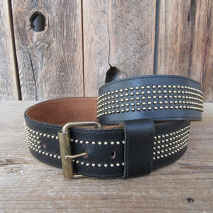 K&ouml;nnte beinhalten: Ein schwarzer Lederg&uuml;rtel mit einer goldenen Schnalle und Reihen von goldenen Nieten. Der G&uuml;rtel ist aus echtem Leder und hat die Worte "GENUINE LEATHER" auf der Innenseite gepr&auml;gt.