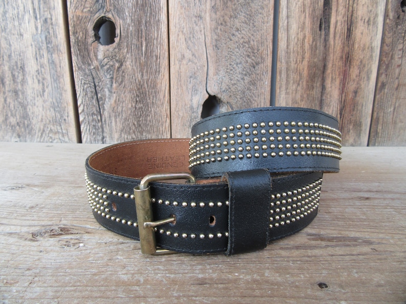 K&ouml;nnte beinhalten: Ein schwarzer Lederg&uuml;rtel mit einer goldenen Schnalle und Reihen von goldenen Nieten. Der G&uuml;rtel ist aus echtem Leder und hat die Worte "GENUINE LEATHER" auf der Innenseite gepr&auml;gt.