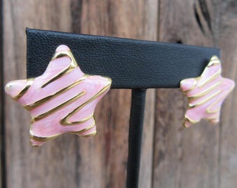 Op de afbeelding: Een paar roze stervormige oorbellen met gouden accenten. De sterren hebben een swirl-patroon en worden getoond op een zwarte oorbellenstandaard.