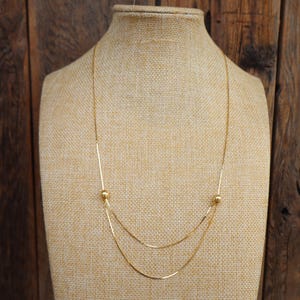 May include: A gold-colored necklace with a layered design. The necklace features two strands of delicate chain, each with a small, round accent. The necklace is displayed on a beige mannequin against a wooden background.