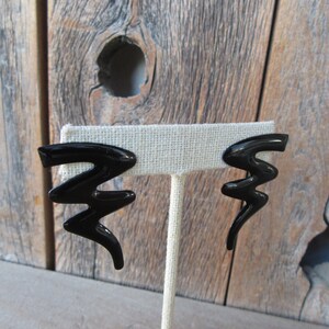 Pode incluir: Um par de brincos pretos de forma abstrata. Os brincos s&atilde;o feitos de um material preto brilhante e t&ecirc;m a forma de um raio.