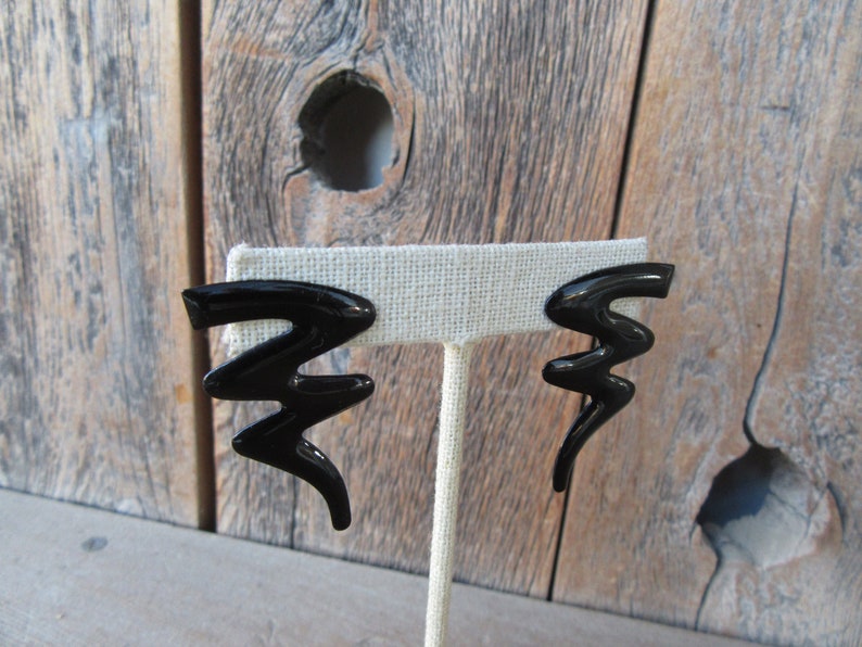 Pode incluir: Um par de brincos pretos de forma abstrata. Os brincos s&atilde;o feitos de um material preto brilhante e t&ecirc;m a forma de um raio.