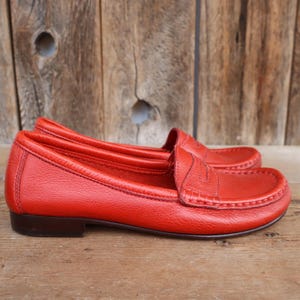 May include: A pair of bright red leather loafers. The shoes have a classic penny loafer design with a stitched vamp and a low heel. The leather has a textured appearance, and the shoes are set against a wooden background.